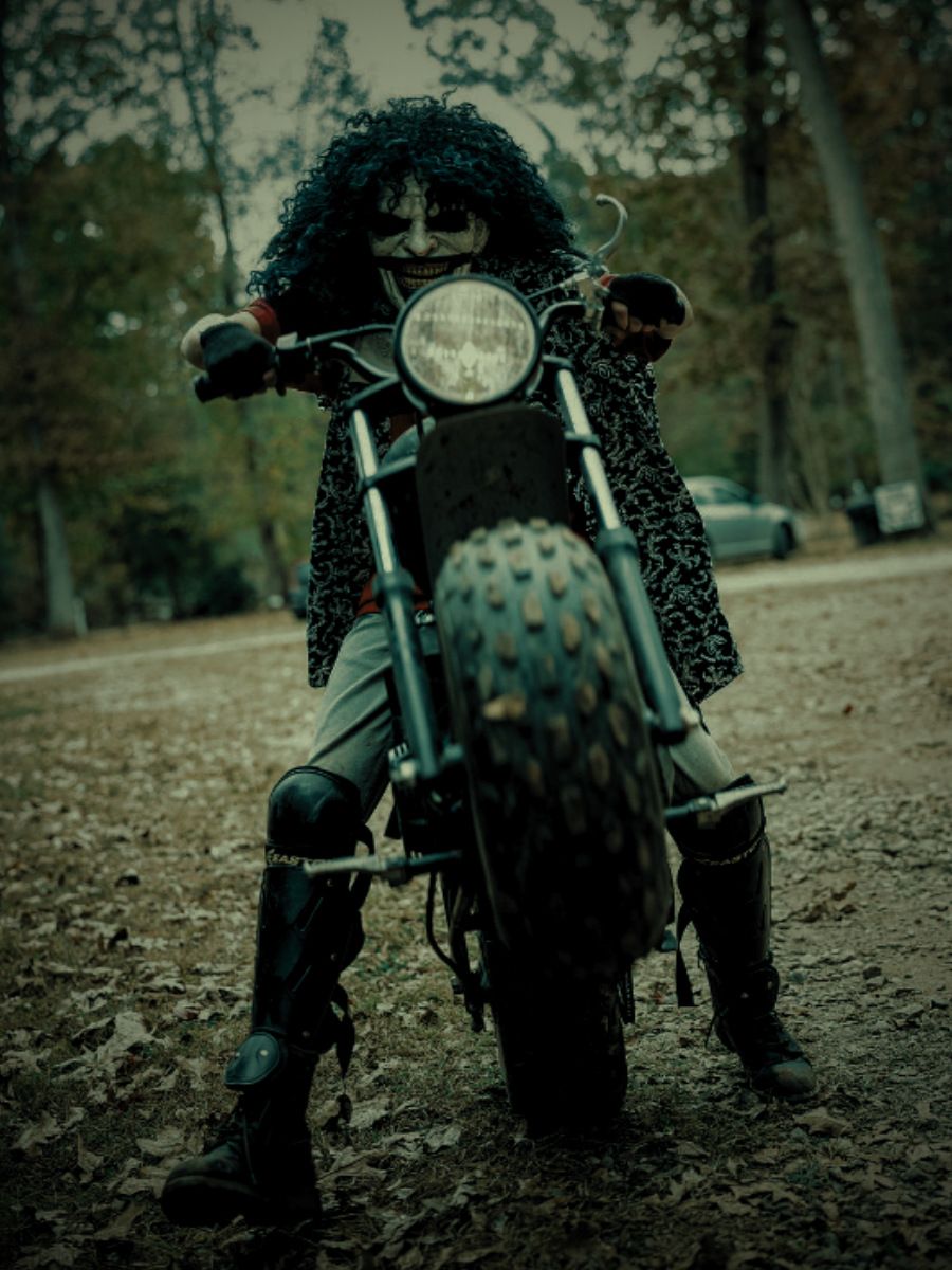 Ashes: a long-haired figure in a leopard-print coat straddling a motorcycle on a leaf-covered road.