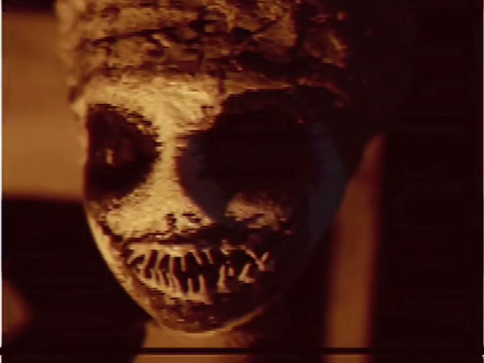 A frame from the trailer: a cracked, hollow-eyed mask lit from below.